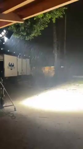 From the shelter of a wooden overhang, a night scene unfolds, illuminated by a powerful spotlight. Rain falls heavily, visible as streaks in the bright light, across an outdoor area that seems to be covered in loose material. In the background, a white truck with a logo resembling a muscular man or mythological figure is parked. Behind the truck, there's a wooden structure, possibly a small building or shed. A light stand with multiple lamps is visible on the left side of the frame. The environment suggests a film set or an area prepared for a night shoot, with the artificial rain enhancing the scene.