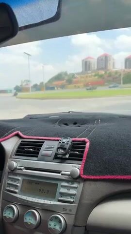 A person is driving a car. The driver's hands are visible on the steering wheel. The radio is playing. There are buildings in the distance. The car is moving on a road. A side mirror view shows a roadside. The windshield is dirty.