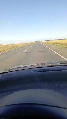 The video shows a first-person view from inside a car looking out through the windshield. The car is driving on a paved road through a flat, open landscape. The road has a white dividing line and appears to be in good condition. On either side of the road are fields of dry grass or crops, stretching to the horizon. The sky is clear and blue, with a few faint clouds. The dashboard of the car is visible in the foreground, and the steering wheel is partially visible at the bottom. The scene suggests a sunny day and a journey through a rural or agricultural area. There are no other vehicles or people visible in the frame.