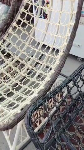 The video displays a close-up, slightly shaky view of several woven hanging chairs. The camera pans across two chairs in the foreground, one with a light-colored weave and the other with a dark weave. The chairs are constructed with a woven material, possibly rattan or wicker, forming a basket-like structure. The background shows a white appliance, possibly a refrigerator, and other furniture items that are out of focus. The environment appears to be an indoor or covered outdoor space, possibly a store or showroom, with a concrete or tiled floor. There are no people visible in the video.