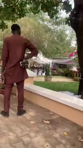 The video shows an outdoor event, possibly a wedding or a party. Men in traditional African attire stand near a white tent where other people are seated at tables. Some children are also present, sitting on plastic chairs. The ground is paved with patterned bricks and scattered with leaves. There are trees and potted plants around, adding to the outdoor ambiance. The camera moves slightly, providing different views of the scene.