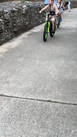 Two young boys are riding bicycles on a concrete path. The boy in the foreground is on a bright green fat-tire bicycle. He is wearing a beige t-shirt, beige shorts, and sandals, with a black bag slung across his chest. The second boy, further ahead and slightly to the right, is on a smaller bicycle, possibly pink and white. He is wearing a white tank top, blue shorts, and blue sandals. The path is bordered on the left by a stone wall constructed with rocks contained in wire cages. In the background, there are some green bushes and parts of a building with a blue roof. The lighting suggests it is daytime.