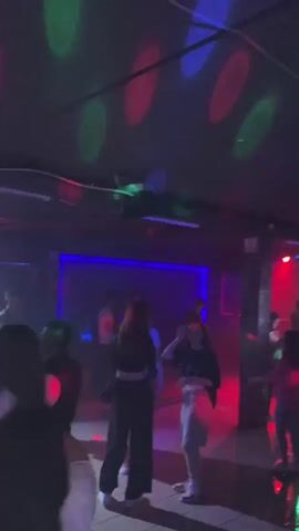 A dimly lit nightclub is filled with colorful laser lights of red, green, and blue projected onto the ceiling and dance floor. Several people are dancing and socializing. A live band is performing on a raised stage at the back of the room, with a keyboardist and two vocalists visible. The overall atmosphere appears to be festive and energetic.