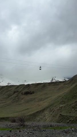 The scene is a mountainous landscape under a cloudy, overcast sky. A small, open-air cable car, appearing as a dark silhouette, hangs suspended from parallel wires strung across the scene. In the background, snow-capped mountains partially obscured by clouds create a wintry contrast to the green and brown hillside in the foreground. A small collection of rocks lies at the bottom of the hill.