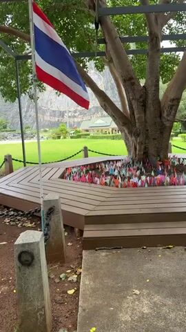A Thai flag waves from a tree to the left of the frame. Below the tree, there is a wooden platform with steps leading up to it. The base of the tree is surrounded by colorful flags stuck into the ground. In the background, a large, intricately carved image is visible on a cliff face. The area is a grassy park with a fence and chain barrier in front of the platform.