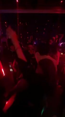 A group of people dancing in a nightclub are captured in a vertical full shot with red lighting. The camera captures a man in the foreground on the left, mostly obscured by another person, while a woman with long hair wearing a black top smiles as she sways to the music, facing right. She is partially covered by a man in a white shirt, who is dancing next to her. A man in a black shirt and long hair stands in the background on the far right, holding a drink and raising his left hand in the air. On the left side of the shot, a person with dark clothing holds a phone up to record the scene. The room is dimly lit with red lights, and additional red vertical lights decorate the ceiling. A band plays in the background, to the left of the man in the white shirt.