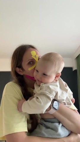 The video shows a woman holding a baby. The woman is wearing a yellow shirt and has yellow and pink tape on her face. The baby is wearing a beige sweatshirt and grey pants. The woman is holding the baby close and kissing it on the head. The baby is looking at the camera and smiling. The room is brightly lit and has a dark grey wall.