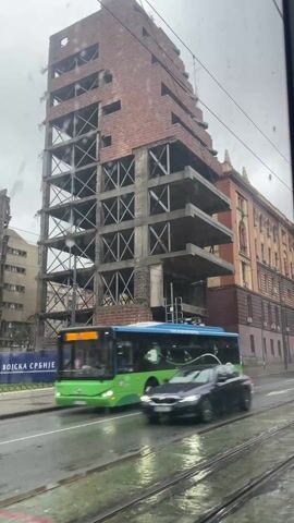 The video shows a view from inside a vehicle, looking out at a street on a cloudy day. Rain is visible on the window. On the left side of the street is a partially constructed building, featuring a brick exterior and exposed concrete and metal supports. The building has a stepped design, getting narrower as it rises. In front of the building are traffic barriers with Serbian text. To the right of the unfinished building is a completed, more traditional-style building with orange and brown exterior. Cars drive by on the wet street, including a black car and a green and blue bus.  A traffic light is visible. The road also features tram lines.