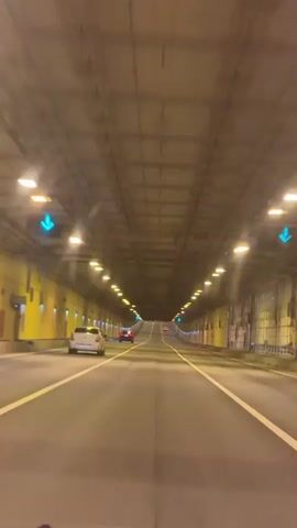 This media file captures a first-person perspective of driving through a tunnel. The scene is characterized by the man-made structure of the tunnel, with a concrete ceiling and walls illuminated by a series of yellow lights. The road ahead is paved and marked with white lines, indicating multiple lanes of traffic. Several vehicles are visible in the distance, including a white car in the left lane and a red car further ahead. Green arrow lights are intermittently visible above the lanes, suggesting traffic flow indicators. The overall atmosphere is one of travel within an enclosed, artificial environment. The time of day and weather conditions are not discernible from the interior of the tunnel. No visible text is present in the media. The location context is identified as Kronstadt, Russia, suggesting this is a tunnel within that area.