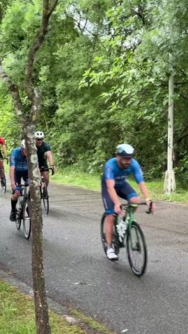 Three cyclists are biking on a paved road, likely during a race or a group ride. The cyclist in the front wears a blue jersey and white helmet. Behind him, there's another cyclist in a green shirt and helmet. Further back is a cyclist in a red jersey and helmet. They're surrounded by lush greenery, with trees lining the side of the road. A tall pole stands near the road. The cyclists are moving at a fast pace, suggesting they're engaged in some sort of competition.