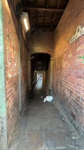The video shows a narrow alleyway with brick walls and a stone floor. The alleyway leads to another opening. A person wearing white shorts, a dark t-shirt, and a white baseball cap with "LUCKY" written on it walks into the alleyway and then towards the camera. Other people are visible in the background near the opening of the alleyway.