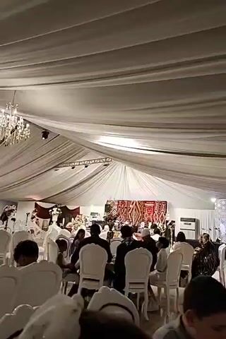 The video shows an indoor event space with a white draped ceiling and a large chandelier. Numerous people are seated at round tables, facing a stage area decorated with a red and gold tapestry. Some attendees are wearing white clothing and head coverings. The lighting is dim, with spotlights illuminating the stage. A large white air conditioning unit is visible on the right side of the stage. The overall atmosphere appears to be that of a formal gathering or celebration.