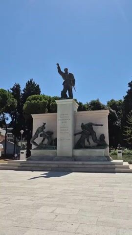 A monument dedicated to the National Liberation Struggle and victims of fascism stands under a clear blue sky. The monument features a central pillar with inscriptions in Croatian, flanked by bronze sculptures depicting scenes of struggle. Atop the pillar, a bronze statue of a standing figure with a raised fist and a sword is visible. Green trees frame the monument in the background. The ground in front of the monument is paved with light-colored stone. The sun casts shadows, indicating a bright, daytime scene.