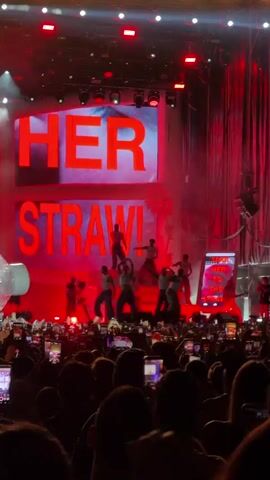 This is a video of a live concert performance. A large stage is illuminated with red lights and features massive LED screens displaying the words "HER" and "STRAW!" in bold red lettering. In front of the screens, a group of performers in matching outfits are dancing. There is smoke or fog on stage, creating a dramatic atmosphere. To the left of the stage, a large, translucent, inflatable object, possibly a bottle or a straw, is visible. To the right, a vertical screen displays the words "HER TO THE STRAW!" in white text. The stage is filled with theatrical lighting. In the foreground, a large crowd of concertgoers is visible, many of them holding up their phones to record the performance. The audience appears to be enthusiastically engaged with the show. The overall impression is a high-energy, visually striking live music event.