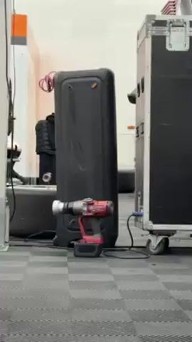 Here is a concise description of the video:

The video shows the environment surrounding a race car in a garage. The scene includes racing equipment such as tires, a drill, and a sound system, all arranged on a checkered floor. The race car, a Porsche, is visible at various angles, highlighting its rear, exhaust, and racing decals. A bald man wearing sunglasses and tattoos is seen lounging in the background, adding a human element to the scene. The setting is a well-lit tent or garage space, suggesting preparation for or aftermath of a race.