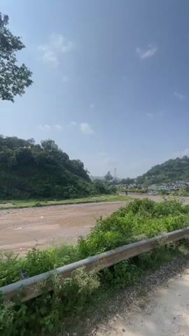 The video shows a view of a dry riverbed with a road passing over it. The landscape is hilly and covered with trees and vegetation. The sky is clear blue with a few scattered clouds. There are some buildings visible on the hillsides. The camera is moving, possibly from a vehicle, giving a sense of travel.