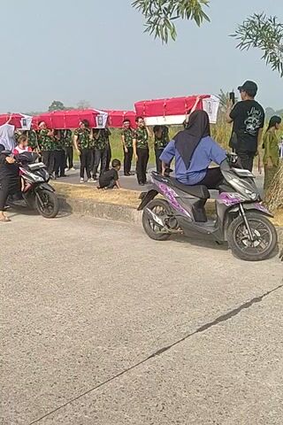 The video appears to be filmed outdoors on a sunny day. A line of people in uniform, possibly students or cadets, stand at attention next to a row of red barriers. Several motorcycles are parked nearby. One person is seated on a motorcycle, wearing a blue hijab and a blue garment. Another person, wearing a black shirt and a black head covering, stands behind them. In the foreground, another motorcycle is visible with two people seated on it. One is wearing a white shirt and a white cap, and the other is wearing a dark shirt and a dark head covering. A third motorcycle is visible in the lower part of the frame with two people on it, one wearing a white shirt and the other a dark shirt and black pants. The overall scene suggests an organized event or gathering.