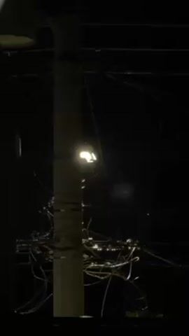 The shot is a nighttime view of a white utility pole with a streetlight attached. Wires are wrapped around the pole and extend out in various directions. The background is dark.