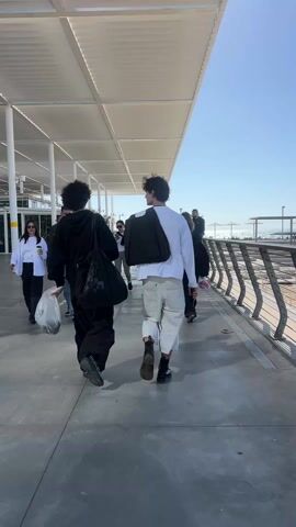 The video shows a group of people walking along a covered walkway near a waterfront. The walkway has a gray concrete floor and is lined with a metal railing on the right side. On the other side of the railing, the ocean can be seen under a clear blue sky.

The main focus is on two people walking away from the camera. The person on the left wears a black hoodie, baggy black pants, and carries a black bag and a plastic bag. The person on the right wears a white long-sleeved shirt, light-colored shorts, and carries a dark jacket over their shoulder.

Other people are walking in the same direction. Some are visible in the background, and some pass by in the opposite direction. There are also people standing by the railing, looking out at the ocean. The walkway is covered by a white, geometrically patterned roof, supported by white columns. The overall environment appears to be a modern, public space, possibly a pier or a promenade.