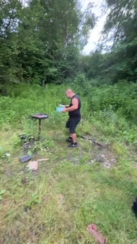 A bald man with a beard, wearing a black tank top and ripped black shorts, stands in a grassy outdoor area surrounded by tall green foliage and trees. He holds a light blue bowl in his left hand and a utensil in his right hand, swaying and dancing to music. To his left is a small, black, round barbecue grill on a stand, with some charred material visible inside. Various small pieces of debris, including cardboard and wood, are scattered on the ground near the grill. A small child, wearing a light blue and yellow patterned hoodie, walks into the frame from the right and briefly approaches the man before turning to face the camera. The man continues to dance.