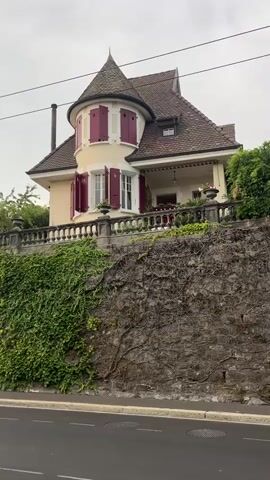 The video opens with a view of a charming, multi-story house with a conical turret and red shutters, situated on a hill. A stone wall covered in lush green ivy separates the house from the street below. The camera then pans left, revealing a street with a trolley bus approaching in the distance. The trolley bus is blue and white and is traveling on a wet road. The sky is overcast and the overall atmosphere appears to be calm and serene. The video also captures a lamppost with its wires stretching across the frame, and a few trees and bushes lining the street. There is a single cyclist visible in the distance, also riding on the street. The architecture of the house suggests a European setting, possibly a suburban or rural area. The presence of ivy and the overcast sky indicate a temperate climate. The video is shot from a static position, suggesting it was filmed from a car or a fixed point on the street.