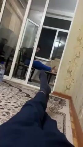 A first-person perspective video shows a person in navy-blue clothing, lying on a patterned rug. To the right, the wallpaper is patterned. In the background, through a set of partially opened glass doors, another person is visible sitting on a chair, looking down, possibly at a phone. A water bottle sits on the floor next to the person on the rug.  The overall lighting is dim.