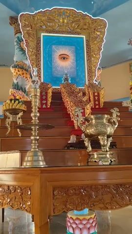 The video shows an ornate altar within a temple or shrine. The altar is tiered and made of wood, with intricate carvings. At the center of the altar, on the highest tier, is a framed image of a single eye glowing with light against a blue background, surrounded by a decorative golden frame. Below this eye image, on a lower tier, are three decorative golden structures with red accents, each displaying Chinese characters. To the left of these structures is a tall, brass-like candelabra with multiple candle holders. To the right, and slightly in front, is a large, ornate golden incense burner with dragon motifs. The background features a ceiling painted with clouds and celestial imagery. The lighting is bright, highlighting the gilded elements of the altar.