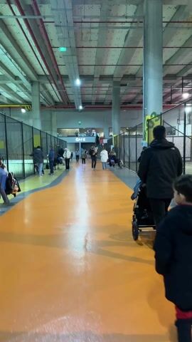 The video shows the interior of a large, open building with high ceilings and concrete pillars. People are walking along an orange walkway, some pushing strollers.  There are fenced-off areas on either side where people are standing or sitting, some appearing to be watching activities in the enclosures. A sign reading "CAFE" is visible in the distance above a stairway. A young woman runs towards the camera along the orange walkway. Overhead, there are exposed pipes and ventilation systems. The overall impression is of an indoor recreational or sports facility.