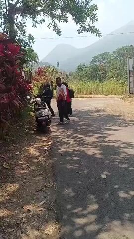 A group of four people are visible on a paved road surrounded by vegetation and a mountainous landscape. A motorcycle is parked on the left side of the road, next to a dense cluster of red-leafed plants. Three individuals are standing near the motorcycle, facing away from the camera. One person, wearing a red hijab and a red long-sleeved shirt, walks away from the group towards the right side of the frame, carrying a green bag. The lighting suggests it is daytime, with shadows cast on the road from overhead foliage. The overall scene appears to be a rural or suburban area on a clear day.