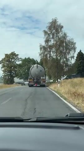 This is a first-person perspective video from inside a vehicle, looking forward. The vehicle is driving on an asphalt road, which appears to be in a rural or semi-rural area. The road has white lines marking the edges. In front of the vehicle, a large silver tanker truck is driving away. The truck has a cylindrical tank with red circular markings and a blue stripe. The rear of the truck is visible, showing its taillights, license plate which reads OL 35 PY, and various pieces of equipment. A pink bucket is hanging from the underside of the truck, near the rear. The environment around the road includes grassy verges, trees, and some buildings in the distance. The sky is cloudy.