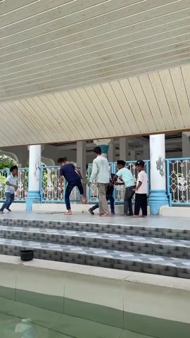 A group of seven boys are playing on a raised platform, possibly a schoolyard or a community gathering space. The platform has a white corrugated roof supported by wooden beams and blue and white columns. A blue metal railing runs along the edge of the platform. The boys are dressed in casual clothing, and most appear to be children or young teenagers. The ground consists of patterned tiles. To the left, there is a body of water, possibly a pool or pond, with some plastic buckets near its edge. The boys are engaged in some form of playful activity, with several of them running or moving in unison. One boy in the center seems to be leading the group. The lighting suggests it is daytime.