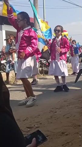 A group of young girls in matching pink and white marching band uniforms are participating in an outdoor parade. One girl in the foreground, on the left, is in motion, her arms extended. Another girl, slightly further back and to the right, holds a flag with green, white, and yellow panels. She is wearing sunglasses. Behind her, other girls in similar uniforms are visible, one holding a flag of similar design. Motorcycles are parked to the left of the parade route, and buildings with red trim are in the background. The ground appears to be a dirt path. The sky is blue and sunny. A hand holding a smartphone is visible in the lower foreground, suggesting the video is being recorded by an observer.