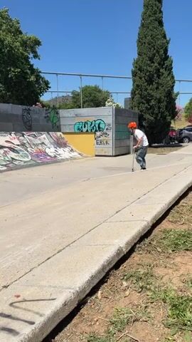 A young person, possibly a male teenager or young adult, wearing an orange helmet, white t-shirt, light blue jeans, and black sneakers, is riding a scooter in a skatepark. The scooter rider approaches a concrete quarter pipe, rides up its curved surface, briefly turns at the top, and then descends back into the flat area of the park.

The skatepark features concrete ramps and walls that are heavily covered in colorful graffiti. Visible text on the walls includes "BMX," "OVER," "Erik," and "FESH," among other illegible tags. In the background, green trees and multi-story buildings can be seen, indicating an urban setting. The sky is clear and blue, suggesting a bright and sunny day, likely midday or early afternoon, as evidenced by the strong shadows cast on the ground and ramps. The overall scene captures an active moment within a vibrant, graffiti-decorated skate environment.