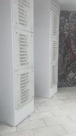 A video shows a memorial with several plaques listing names and historical information. The plaques are mounted on white pillars. One pillar on the left has multiple name lists, each starting with a surname and ending with initials. Another pillar in the center also has name lists. To the right of the center pillar is a mosaic depicting a group of people, likely soldiers, in a battle scene. The mosaic uses a variety of colors, including red, brown, black, and white. A third pillar on the right has a title that appears to be "СЕНТЯБРЬ" (September) and lists numbers of soldiers, units, and casualties. The floor is tiled with large white squares. The lighting suggests an indoor or covered outdoor space with a skylight or open ceiling. The overall impression is of a solemn memorial site.