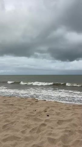 This media file depicts a cloudy day at a beach in João Pessoa, Brazil. The foreground is dominated by textured sand, with a few scattered dark specks, possibly small stones or debris. Beyond the sand, the ocean stretches out, its surface agitated by moderate waves that are breaking and forming white foam as they approach the shore. The water itself appears a muted, greenish-gray.

The sky is filled with heavy, dark gray clouds, suggesting overcast conditions and possibly the threat of rain. The horizon line is clearly defined, separating the sea from the imposing cloudscape. In the mid-ground of the ocean, a few people are visible wading or swimming in the water. One individual is wearing a bright pink garment, standing out against the darker tones of the sea and sky. Another figure is also visible, partially obscured by the waves.

The overall mood evoked by the scene is one of subdued natural beauty, with the dramatic sky and the active ocean creating a sense of raw, untamed nature. The presence of people enjoying the water suggests resilience or a disregard for the impending weather. The time of day is difficult to determine precisely due to the overcast conditions, but it appears to be daytime. There is no visible text in the media file.