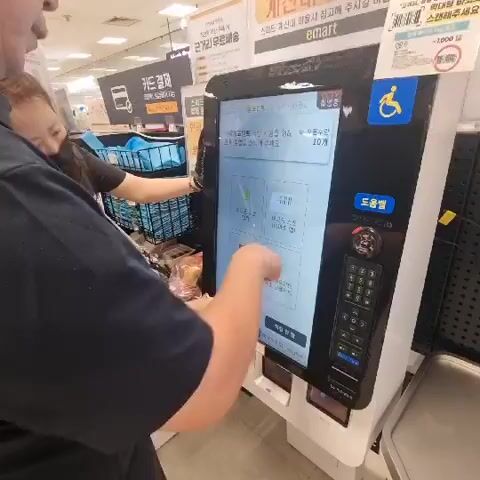 The video shows a man and a woman interacting with a self-checkout machine in what appears to be a supermarket. The man, wearing a dark blue shirt, is in the foreground and is inserting a credit card into the machine. A woman, wearing a black shirt and a mask, is standing next to him and appears to be assisting him. The self-checkout machine has a large touchscreen display showing Korean text and graphics, a keypad, and a card reader slot that is illuminated with a green light. There are also shelves with products visible in the background. The scene is well-lit, suggesting an indoor commercial environment.