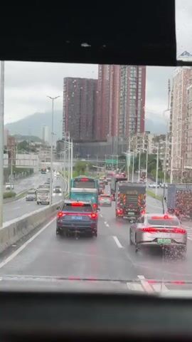 The video shows a view from inside a vehicle, looking out at a busy highway during what appears to be a rainy day. The road is wet, and raindrops are visible on the windshield. Multiple lanes of traffic are moving, with cars, buses, and trucks. In the foreground, a dark-colored car with its taillights illuminated is in the left lane. To its right, a silver sedan is in the middle lane. A large blue bus is in the left lane further ahead. Several trucks, some appearing to be carrying cargo, are also present in the traffic. In the background, tall apartment buildings with red brick exteriors are visible, as are trees and streetlights. The sky is overcast. The traffic appears to be moving slowly, suggesting a potential traffic jam.
