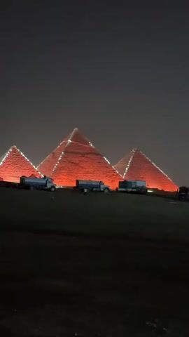 The video depicts the Great Pyramids of Giza illuminated in red light at night. There are several trucks parked in the foreground. The scene is dark, with the sky also appearing dark and hazy.