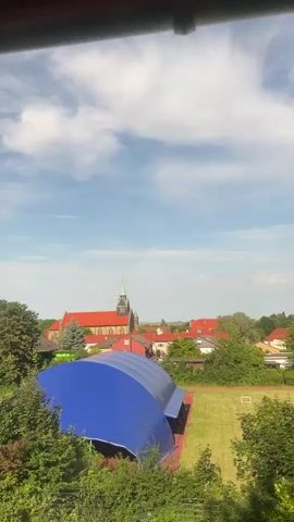 The video captures a serene landscape under a partly cloudy blue sky. In the foreground, lush green trees and bushes partially obscure the view. Dominating the mid-ground is a large, arched structure covered with a bright blue fabric, likely a sports hall or similar facility. Adjacent to it is a well-maintained green field, possibly a sports field, with faint markings and a small white goalpost visible.

Further in the background, a quaint village unfolds. The focal point of the village is a church with a distinctive red roof and a tall, green spire. Several houses with terracotta roofs are scattered amongst the greenery. The scene is bathed in natural daylight, suggesting it's daytime. There are no people or animals visible in the video, and no discernible actions or interactions are taking place. The overall impression is one of a peaceful rural or suburban setting on a pleasant day.