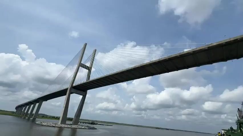 The video shows a wide shot of a large cable-stayed bridge with two tall towers, viewed from below and from the side. The bridge spans a body of water. Partially visible in the foreground are the cranes and deck of a large cargo ship. The sky is partly cloudy with blue patches. The camera appears to be moving sideways, showing different angles of the bridge and the ship. The perspective is low, looking up at the massive structure of the bridge and the ship.