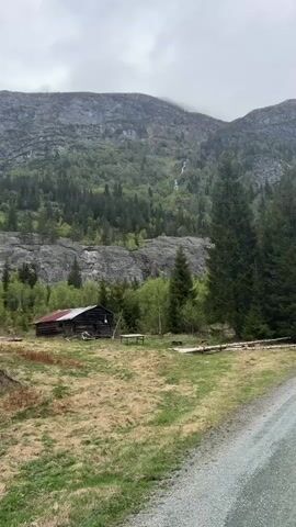The video presents a scenic view of a mountain landscape, featuring a small, old wooden cabin with a red roof in the foreground. Behind the cabin, a rugged mountain covered with green trees and rocky patches rises into the cloudy sky. A thin waterfall cascades down the mountain, visible amidst the foliage. In the lower right of the frame, a gravel road is visible, indicating a path leading to or from the cabin. The overall environment is green and lush, typical of a natural setting, suggesting a rural or wilderness area.