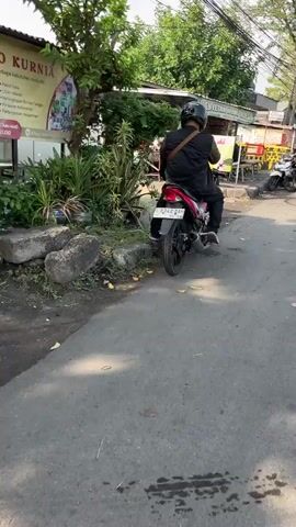 A person wearing a black helmet and a black vest is sitting on a red motorcycle. The motorcycle is parked on the side of a paved road, with its rear wheel facing the camera. The person is looking away from the camera and appears to be interacting with something off-screen. The background shows a street scene with trees, buildings, and other motorcycles parked. There is a yellow sign with text in an unknown language to the left of the motorcycle. The ground is asphalt, and there are some rocks and plants lining the side of the road. The lighting suggests it is daytime.