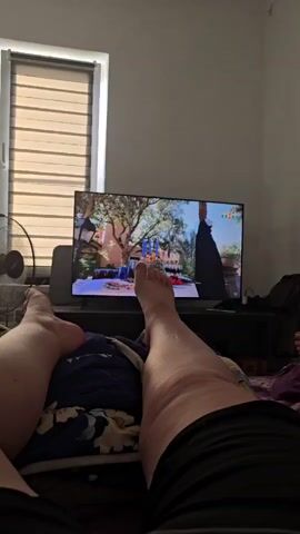 This media file shows a first-person view of someone lying on a bed, facing a television screen. The television displays a scene that appears to be from a television show or movie. The room is dimly lit, suggesting it might be nighttime or the blinds are drawn to create a darker atmosphere for watching.

**Subjects:**
*   **Person:** The legs and feet of a person are prominently in the foreground, indicating the viewer's perspective. The person is lying down, likely on a bed, covered by blankets.
*   **Objects:** A television set is the central object in the scene, displaying various images. A fan is visible to the left of the television. There are also what appear to be blinds on a window behind the television. A dark cabinet or stand is beneath the television.
*   **Scene:** The television screen shows different settings. Initially, it depicts an outdoor scene with trees and a building. Later, it shows a still life with candles and fruit. Finally, it shows an outdoor area with a fountain, plants, and a flag with a face on it.
*   **Setting:** The setting is an indoor room, likely a bedroom, with a bed and a television. The presence of a window with blinds suggests an interior space. The location context provided is Abinsk, Russia.

**Activity or Event:**
The primary activity is watching television. The viewer is positioned comfortably on a bed, engrossed in the program.

**Notable Details:**
*   **Time of Day:** The dim lighting suggests it could be evening or night, or that the room's lighting is intentionally subdued for watching TV.
*   **Location Cues:** While specific visual cues are limited, the provided location context is Abinsk, Russia. The interior furnishings are relatively generic and do not offer strong regional indicators.
*   **Emotions:** The relaxed posture of the person's legs and feet suggests a sense of comfort and leisure.

**Visible Text:**
On the television screen, the logo "THT" is visible at the top right corner during one of the scene changes.