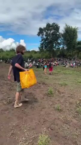 The video shows two men walking through a muddy field full of parked motorbikes and cars. The first man has curly hair and is wearing a dark t-shirt, cargo shorts, and hiking boots, and he carries a large yellow bag. The second man is wearing a black t-shirt, khaki shorts, and a patterned crossbody bag, and he carries a net bag. The ground is muddy and rutted. In the background, there are more motorbikes and cars parked, and a group of people can be seen walking. There are trees and greenery in the distance, and the sky is blue with white clouds.