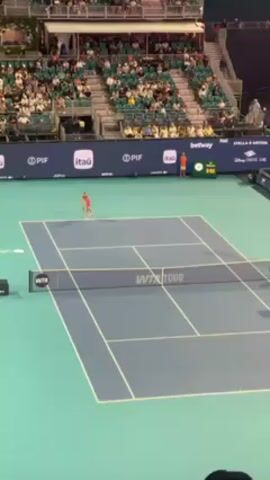 A tennis match is in progress at an outdoor arena with green and blue courts and partially filled stadium seating. A player in a bright pink outfit serves the ball across the net. Behind the tennis court are advertisements and sponsor logos. A digital scoreboard displays the score. The audience is mostly dressed casually. Two officials stand at the back of the court. The scene is shot from a slightly elevated angle.