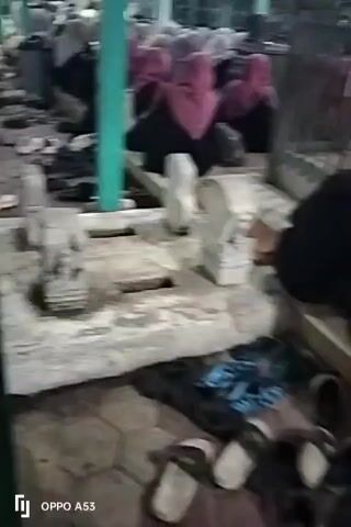 The video shows a group of people, primarily women wearing headscarves, sitting on the ground in an outdoor area. They appear to be engaged in prayer or a religious ceremony. Shoes are scattered around them on a hexagonal tiled ground. There are some structures in the background that suggest a place of worship or a communal gathering space, possibly a mosque or a shrine, with pillars and a green roof visible. In the foreground, there are some grave-like structures made of concrete. The overall atmosphere seems solemn and respectful.