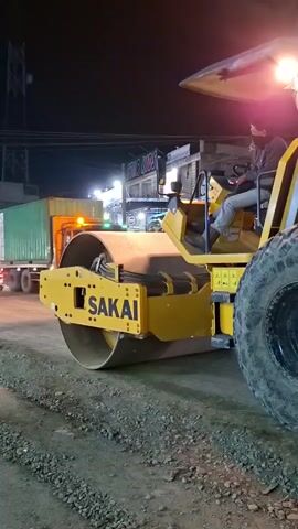 The media shows a yellow Sakai SV526D roller truck being operated by a person. The truck is on a gravel road at night. The roller is the main focus of the image, with its large drum in the foreground. The driver is visible in the cab, wearing a dark shirt and a mask. The scene appears to be a construction site or road maintenance area.

Notable details include:
- Time of day: Night, as indicated by the dark sky and artificial lighting.
- Weather: Not directly visible, but the lighting suggests it is not raining heavily.
- Location cues: The presence of construction equipment and the gravel road suggest an outdoor industrial or infrastructure development setting. The text "Blangpeuria, Indonesia" is provided as context.
- Visible text: "SAKAI" is clearly visible on the roller drum, and "SV526D" and "ECO MODE" are visible on the side of the truck.

The activity taking place is the operation of the roller truck, likely for compacting the road surface. The driver appears focused on their task. There are no strong indications of emotion, but the overall atmosphere is one of work being done at night. The lighting from the truck's headlights illuminates the immediate area.