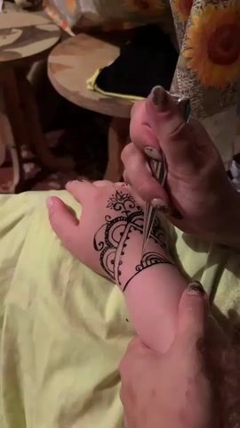 A close-up shot shows a person applying henna to another person's hand. The recipient's hand is resting on a light green fabric. A hand with dark nail polish holds a henna applicator, a conical tool filled with dark henna paste, and is carefully drawing intricate patterns on the back of the recipient's hand and wrist. The henna design appears to be a complex mandala-like pattern with floral elements. In the background, a wooden table and a sunflower-patterned fabric are visible. The lighting is warm, suggesting an indoor setting.