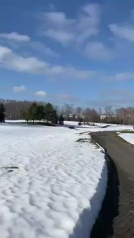 The camera pans from left to right, capturing a bright, sunny day with a partially snow-covered landscape. A brick-paved pathway curves through the scene. To the left of the path, there is snow, and some green grass peeks through.  A small cluster of pine trees stands near the left edge of the frame.  In the distance, a line of deciduous trees is visible.  The sky is blue with scattered white clouds.