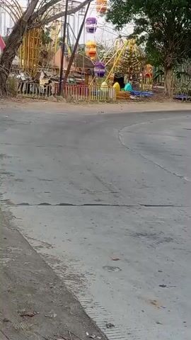 The video shows a street scene with a Ferris wheel in the background. A motorcycle is seen riding on the road. The Ferris wheel appears to be part of a temporary carnival or amusement park, with colorful gondolas. The environment suggests an outdoor setting, possibly in a town or city.